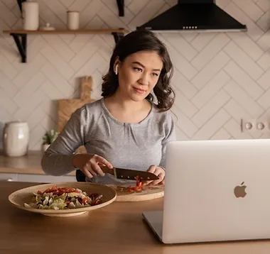 Young woman sitting at kitchen table looking at laptop while slicing vegetables